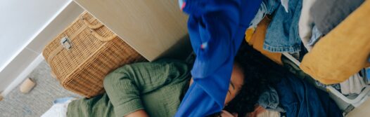 A woman getting covered by her own clothes, as she lays under wardrobe pulling out items.