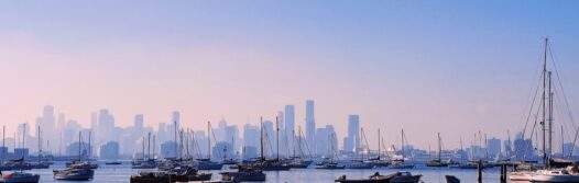 A view of the Melbourne skyline from the Williamstown bay, with boats floating in the water.